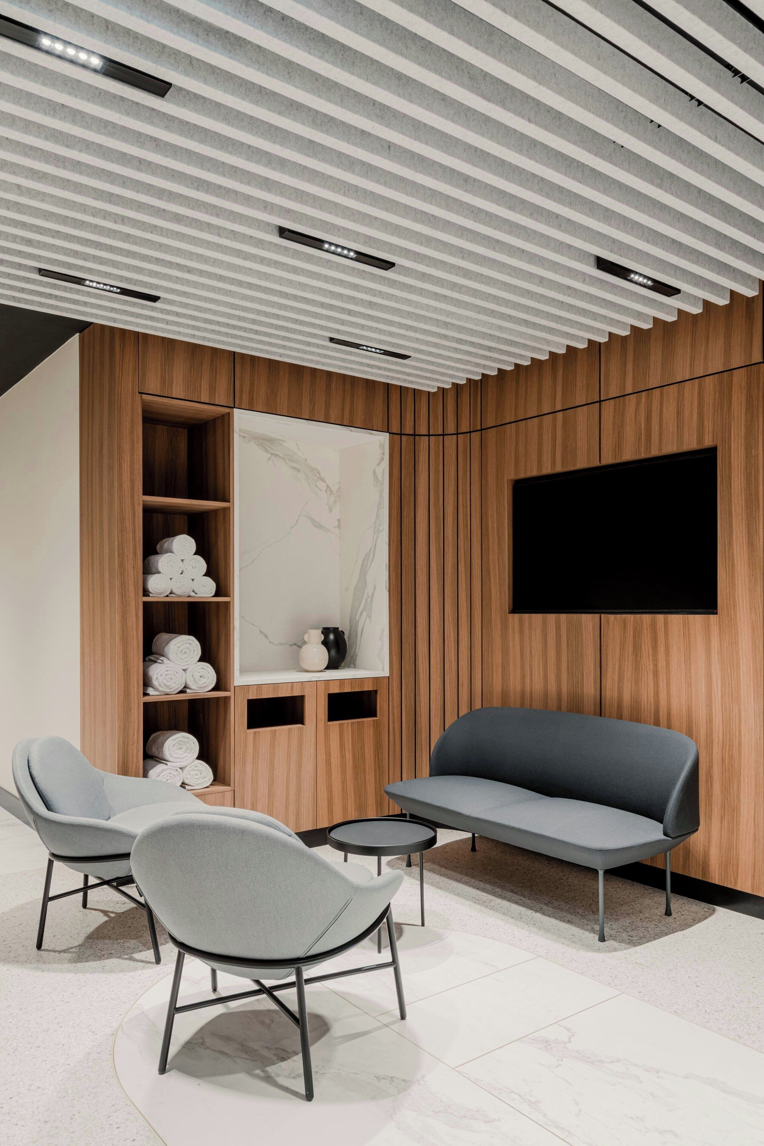 A modern living room with a sofa, two armchairs, a coffee table, and a TV mounted on a wooden wall. Shelves with rolled towels are visible.