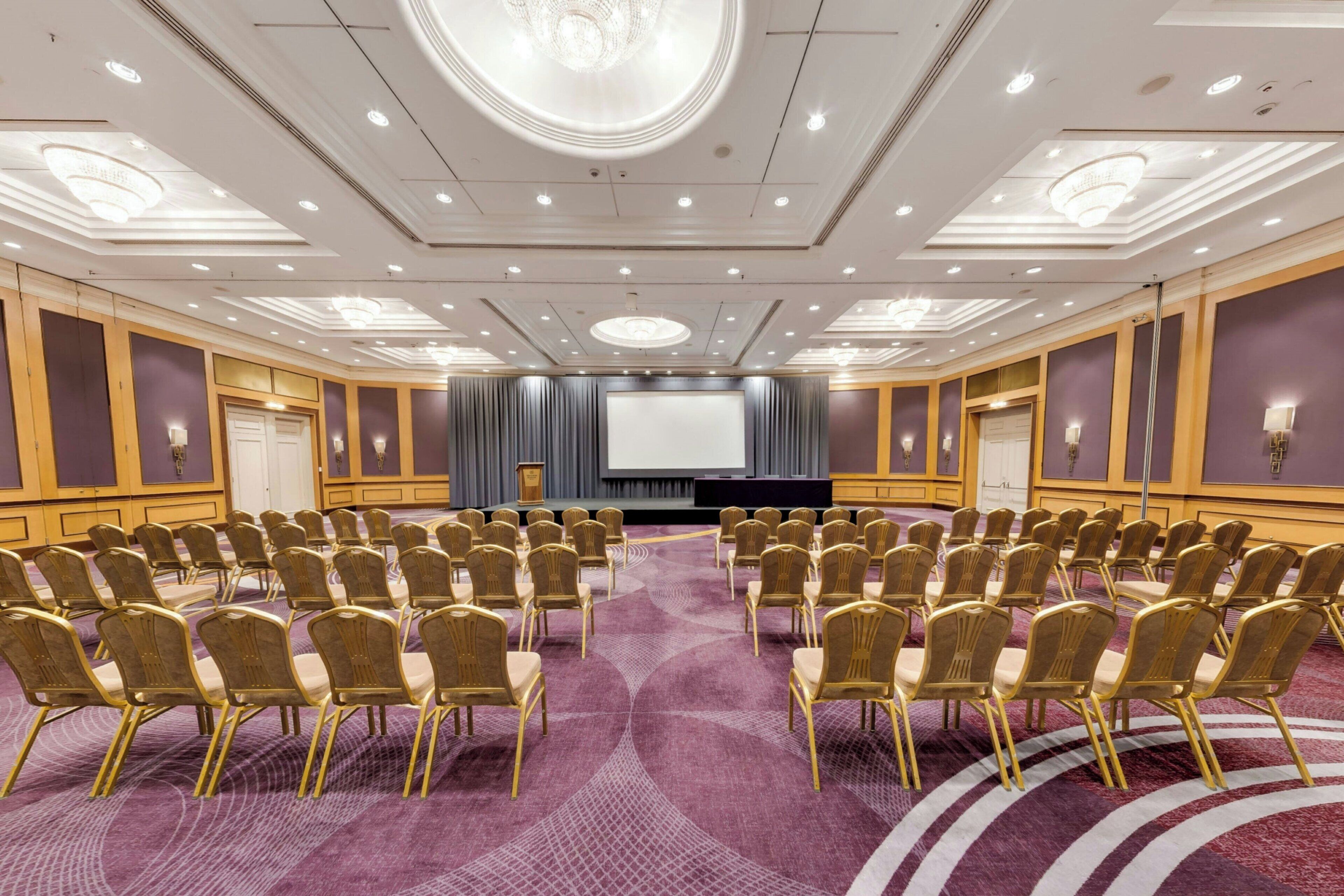 A conference room with rows of gold chairs, a large screen, and a stage.