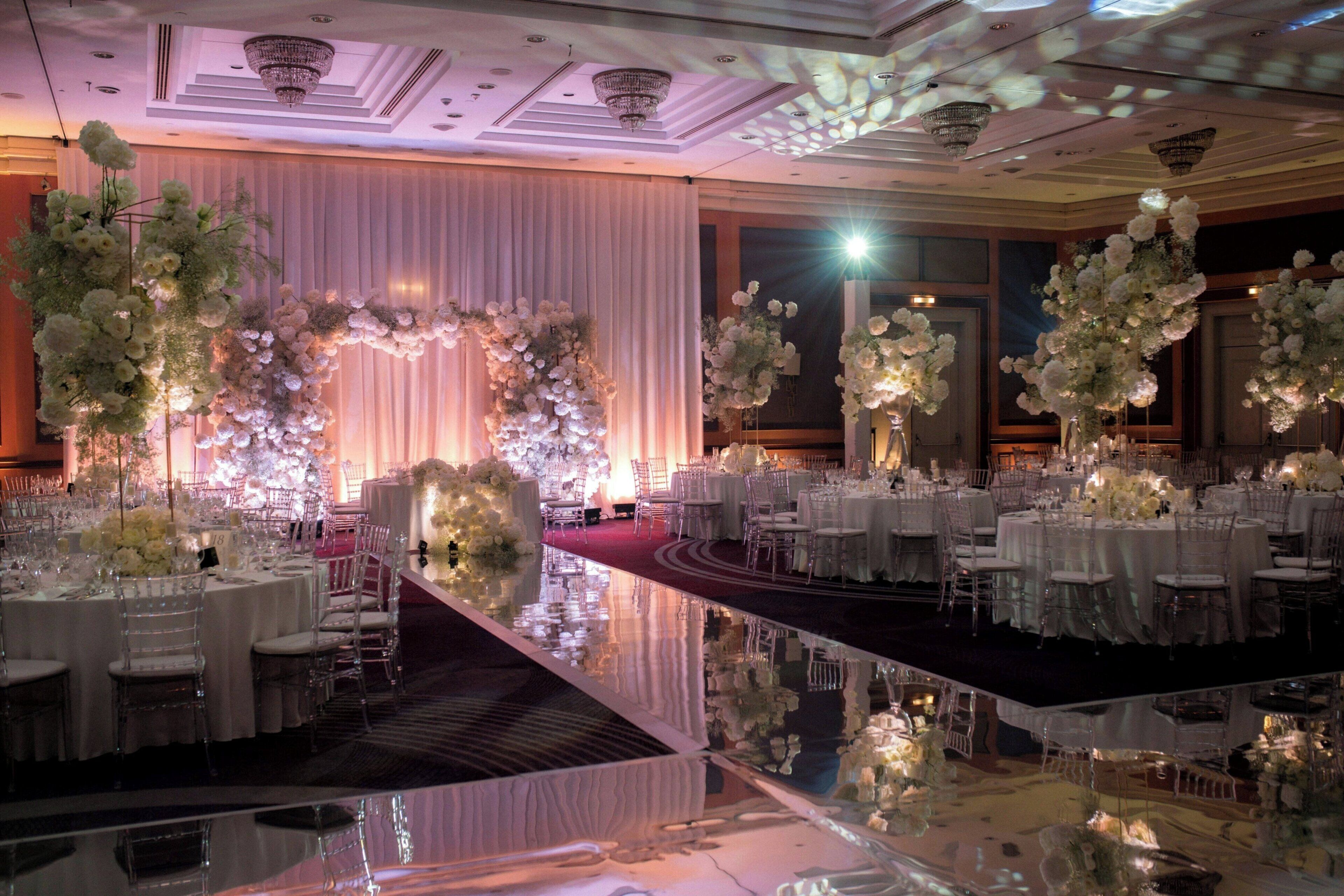 Un salón de banquetes con mesas redondas, arreglos florales blancos y un suelo reflectante.