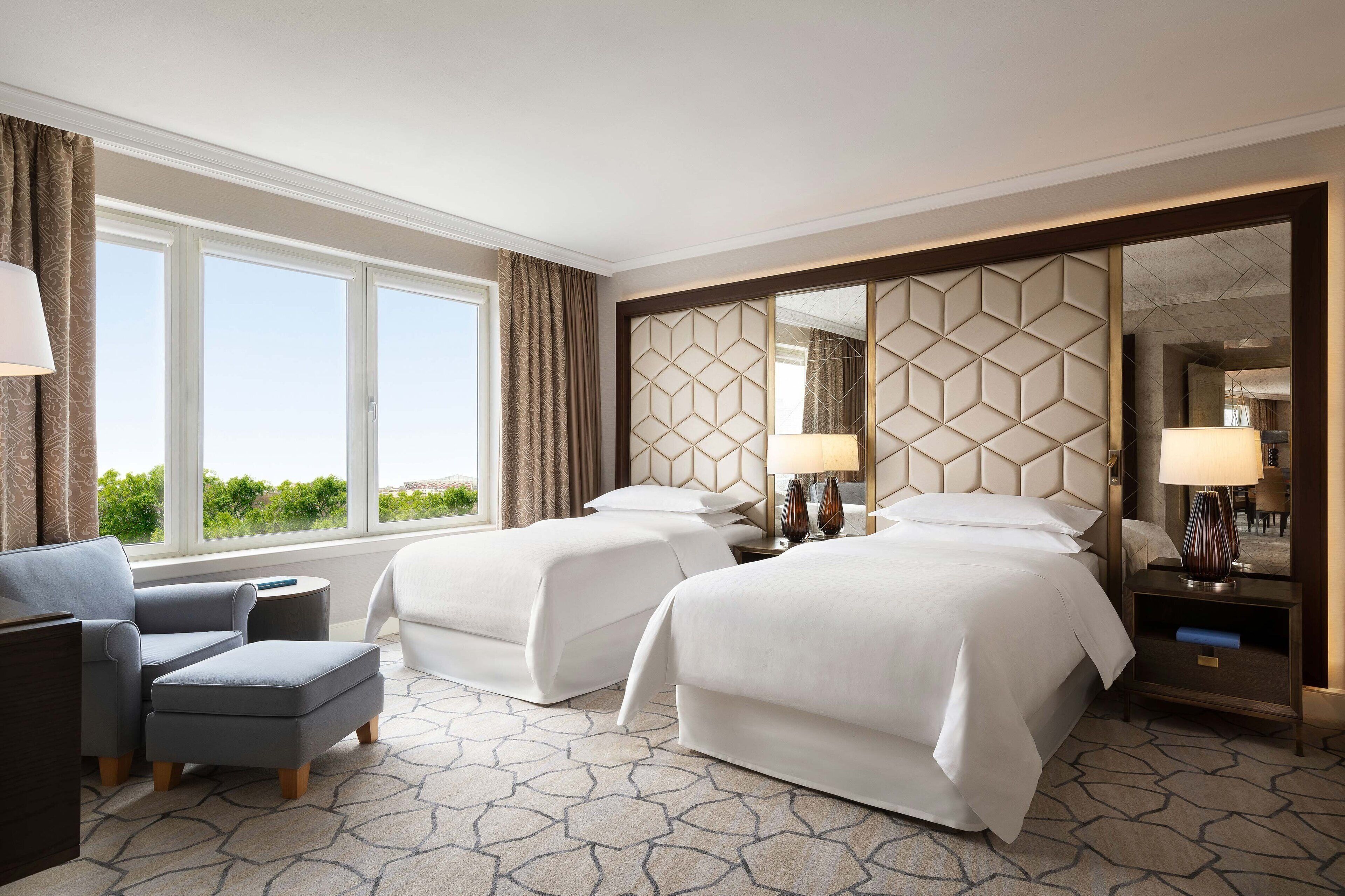 A hotel room with two beds, a sofa, a small table, and a large window with curtains.