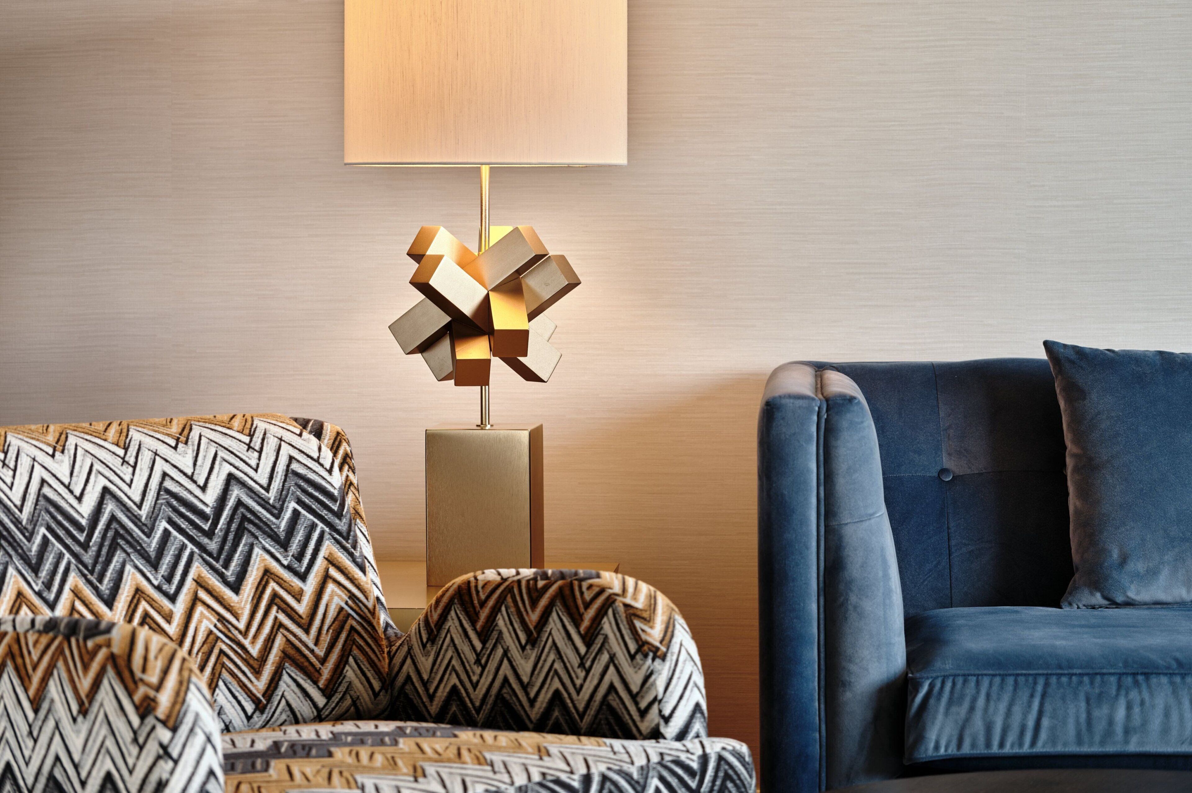 Ein modernes Wohnzimmer mit einer Couch und einem Sessel, einer Lampe mit geometrischem Fuß sowie einer Wandleuchte.