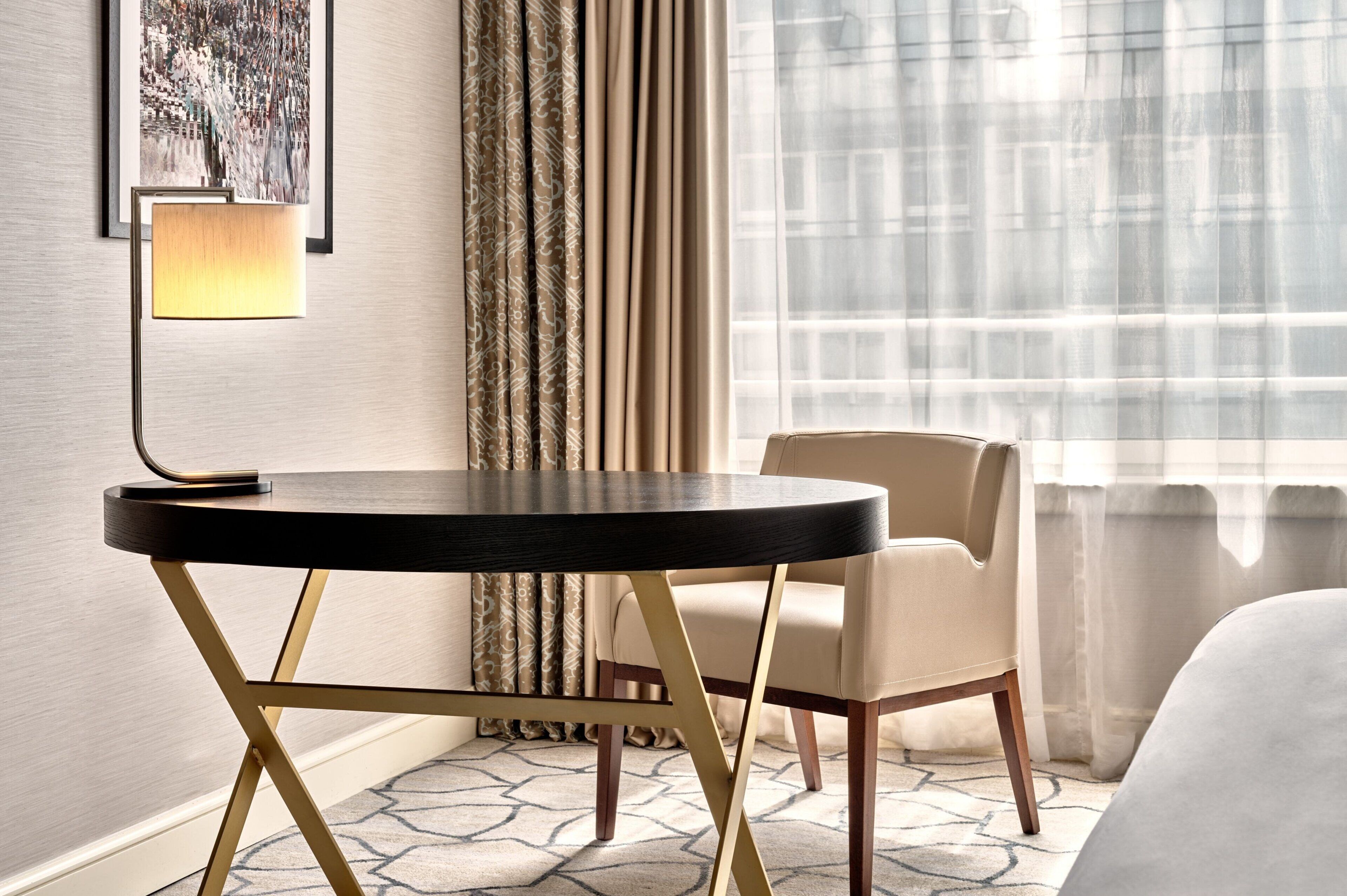 A hotel room with a round table, a chair, a lamp, a patterned rug, and a window with sheer curtains.