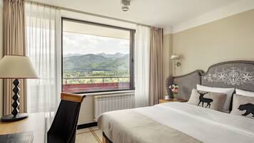Habitación de hotel con una cama grande, un escritorio con lámpara, una silla y vistas a las montañas a través de la ventana.