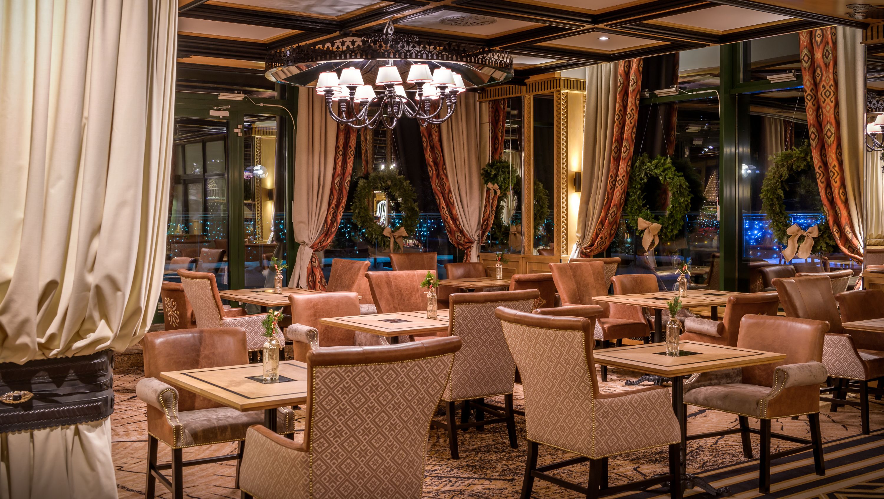 A restaurant with multiple tables and chairs, large windows with curtains, and a chandelier.