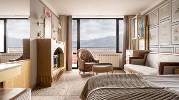 A hotel room with a bed, a sofa, a chair, a fireplace, and large windows offering a mountain view.