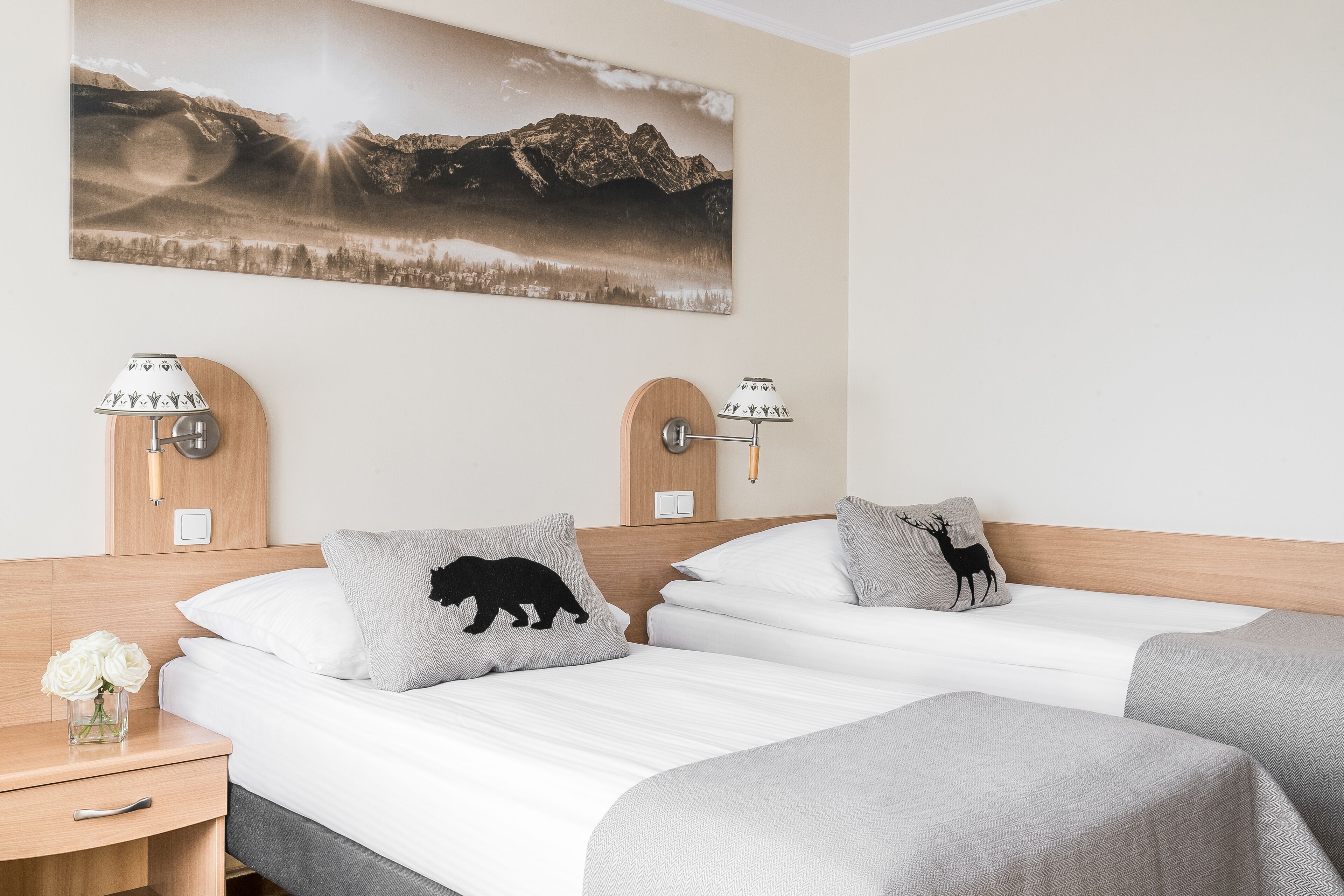A hotel room with two beds, a nightstand with a lamp, a vase with flowers, and a wall-mounted picture of a mountain landscape.