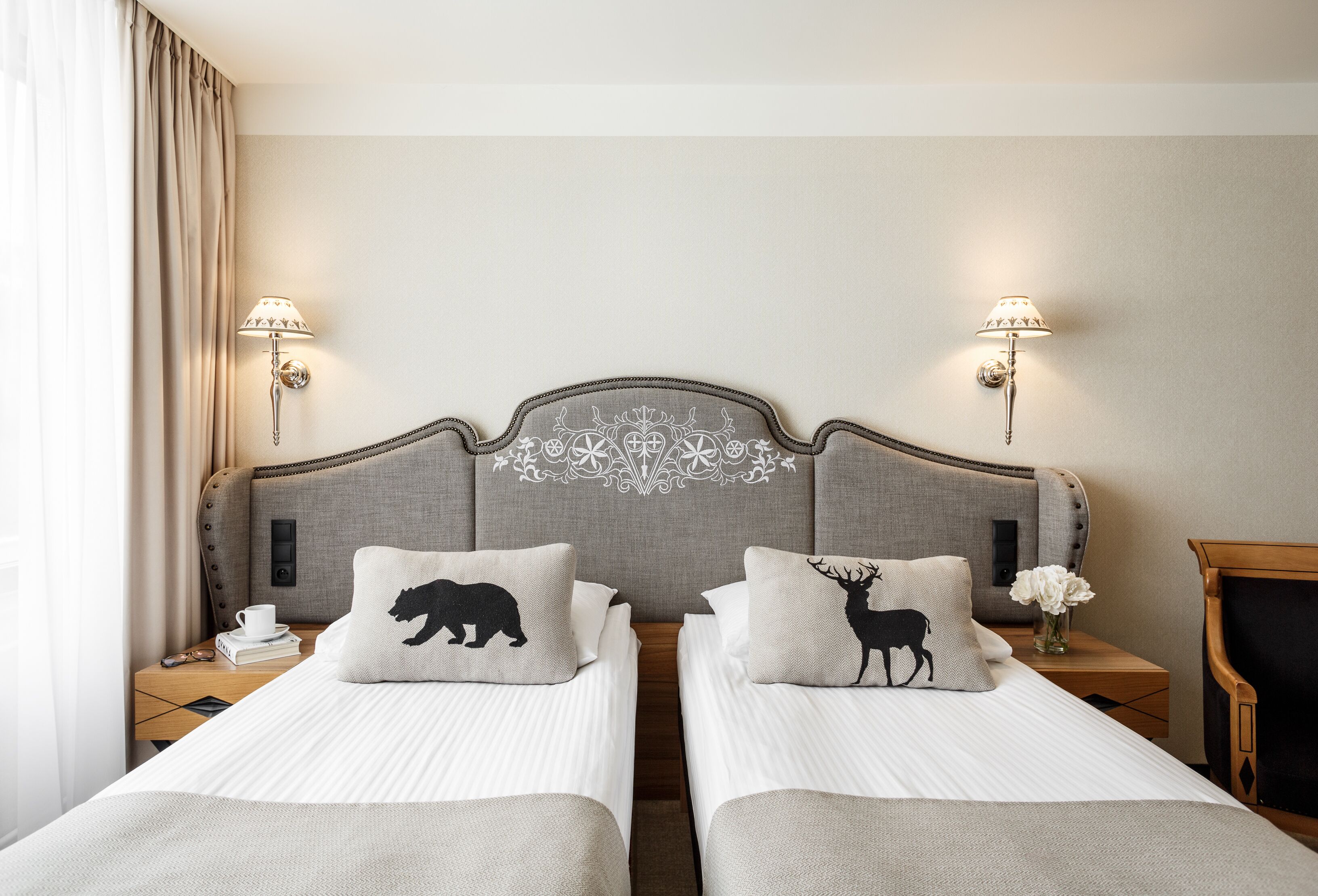 Habitación de hotel con dos camas, cabecero con motivo decorativo, lámparas de noche, mesita de noche con una taza y un libro, y una silla de madera.