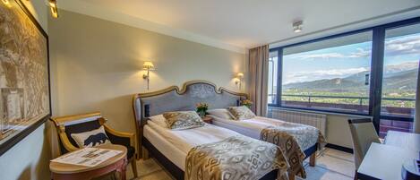 Quarto de hotel com duas camas, uma escrivaninha e uma cadeira. Há uma janela grande com vista para as montanhas.