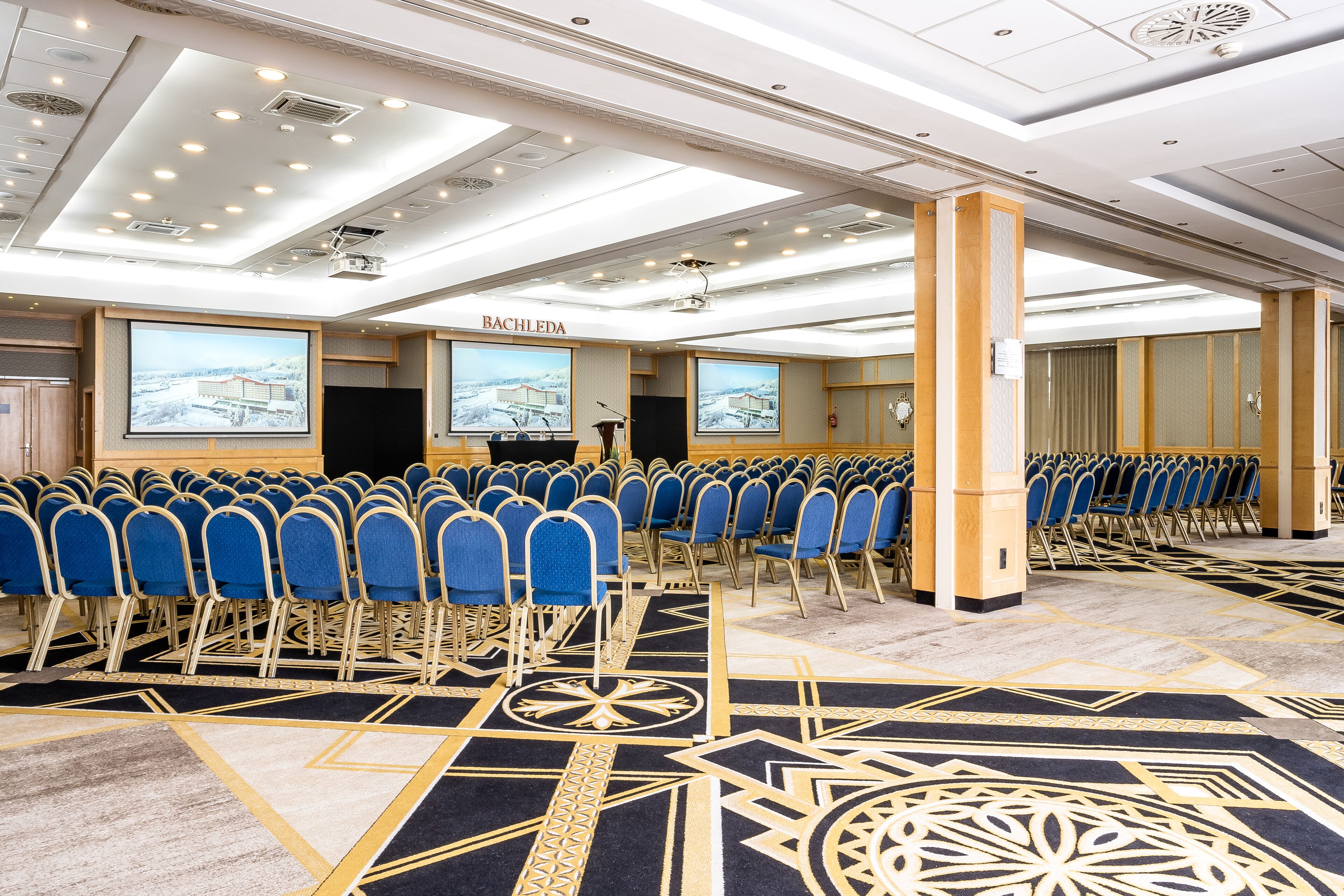 A conference room with rows of blue chairs, a stage with a podium, and large screens displaying a snowy landscape.