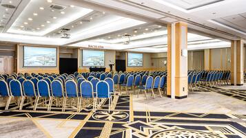 A conference room with rows of blue chairs, a stage with a podium, and large screens displaying a snowy landscape.