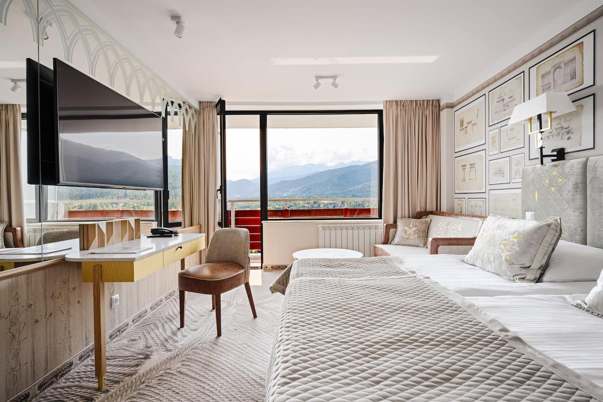 A hotel room with a large bed, a desk with a chair, a flat-screen TV, and a view of mountains through the window.