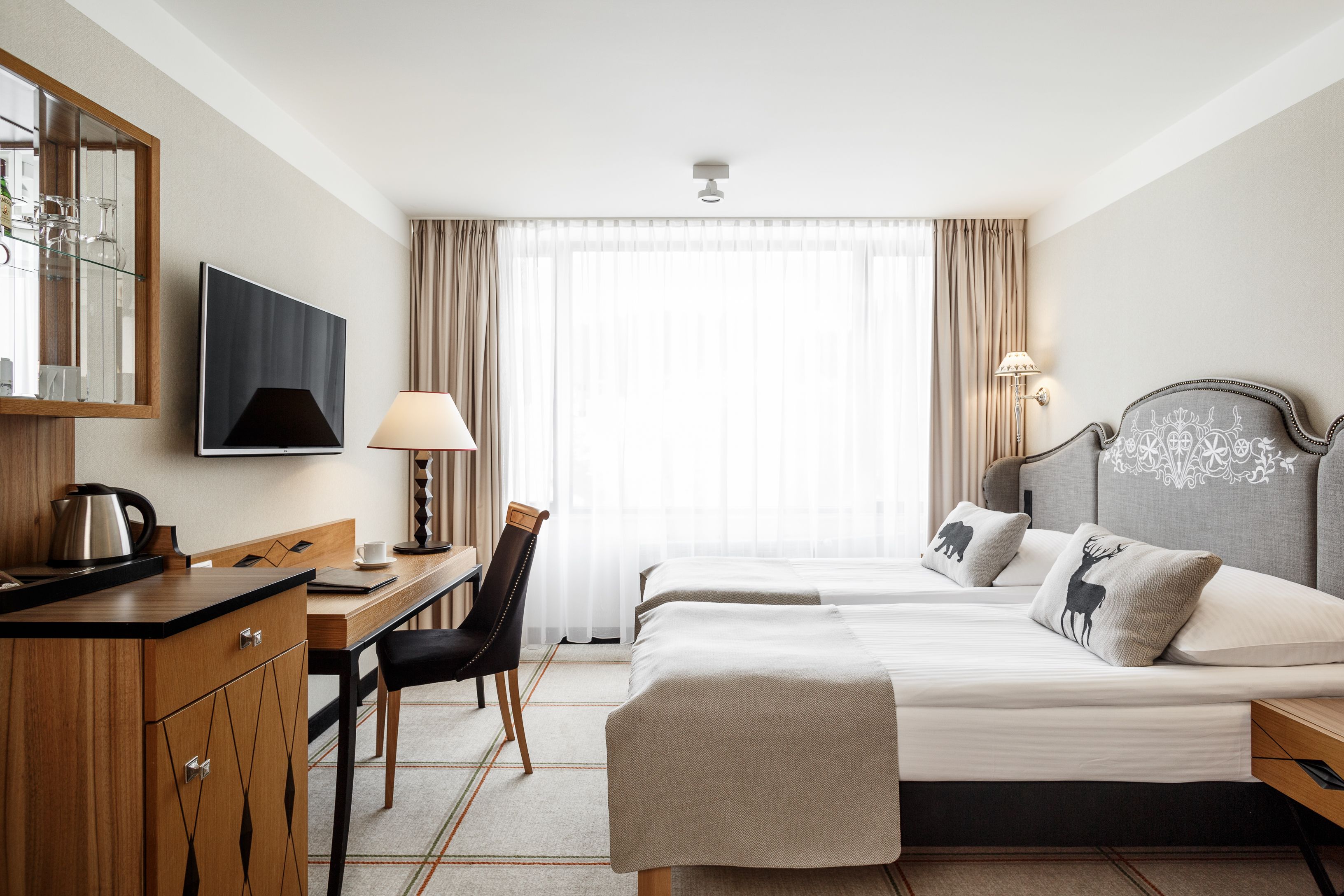 A hotel room with two beds, a desk, a chair, a television, and a window with curtains.