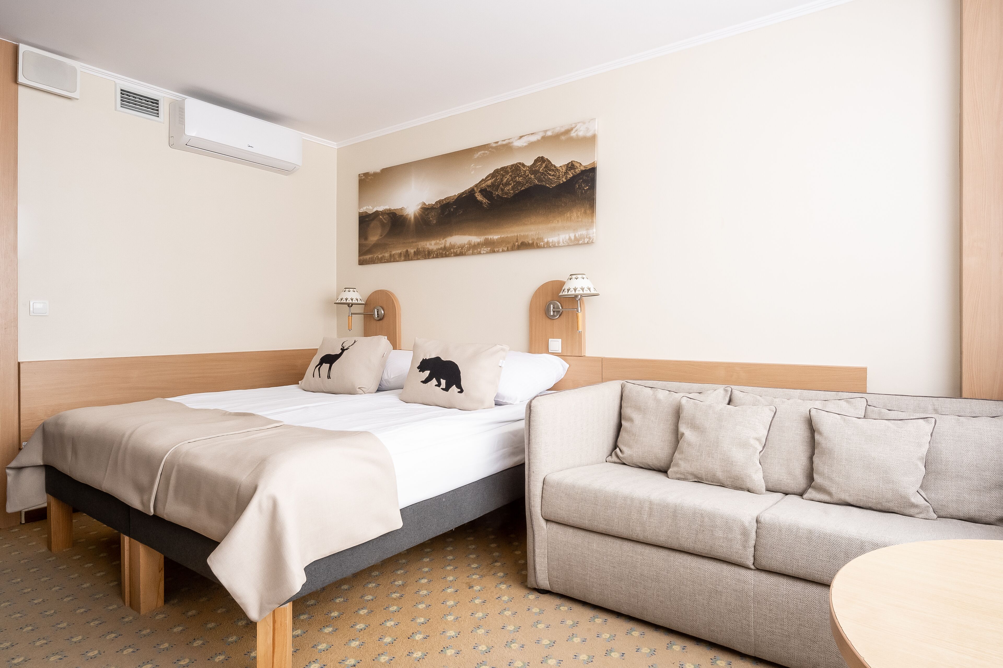 Habitación de hotel con cama, sofá, aire acondicionado de pared, dos lámparas de pared y un cuadro con paisaje de montaña.