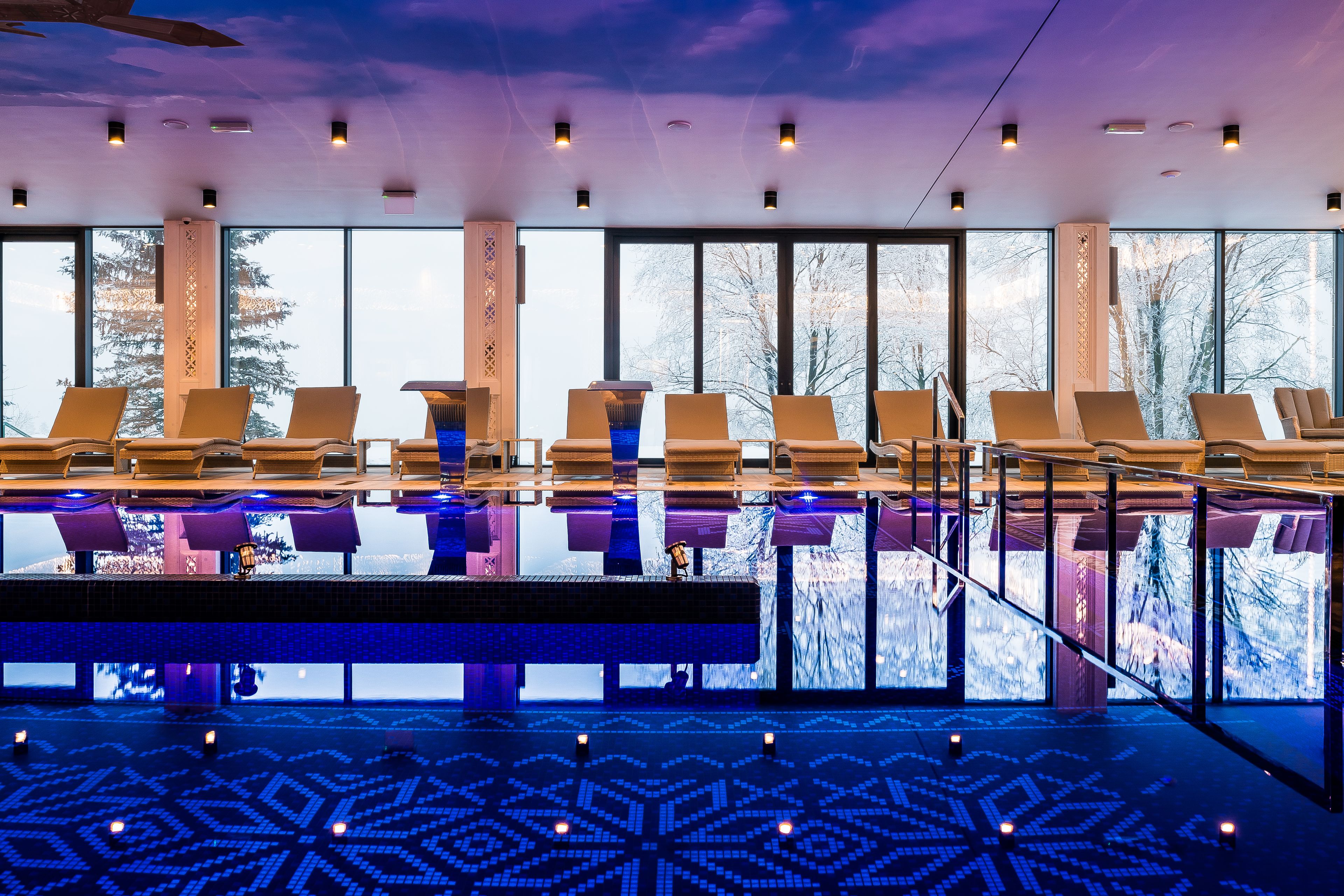 Un espace piscine intérieur moderne, doté de chaises longues et de grandes fenêtres offrant une vue sur le paysage environnant.