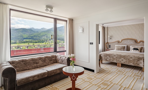 A hotel room with a large window offering a mountain view, a sofa, a small table with a vase, and a bed with patterned bedding. - Bachleda Hotel Kasprowy**** (Zakopane)