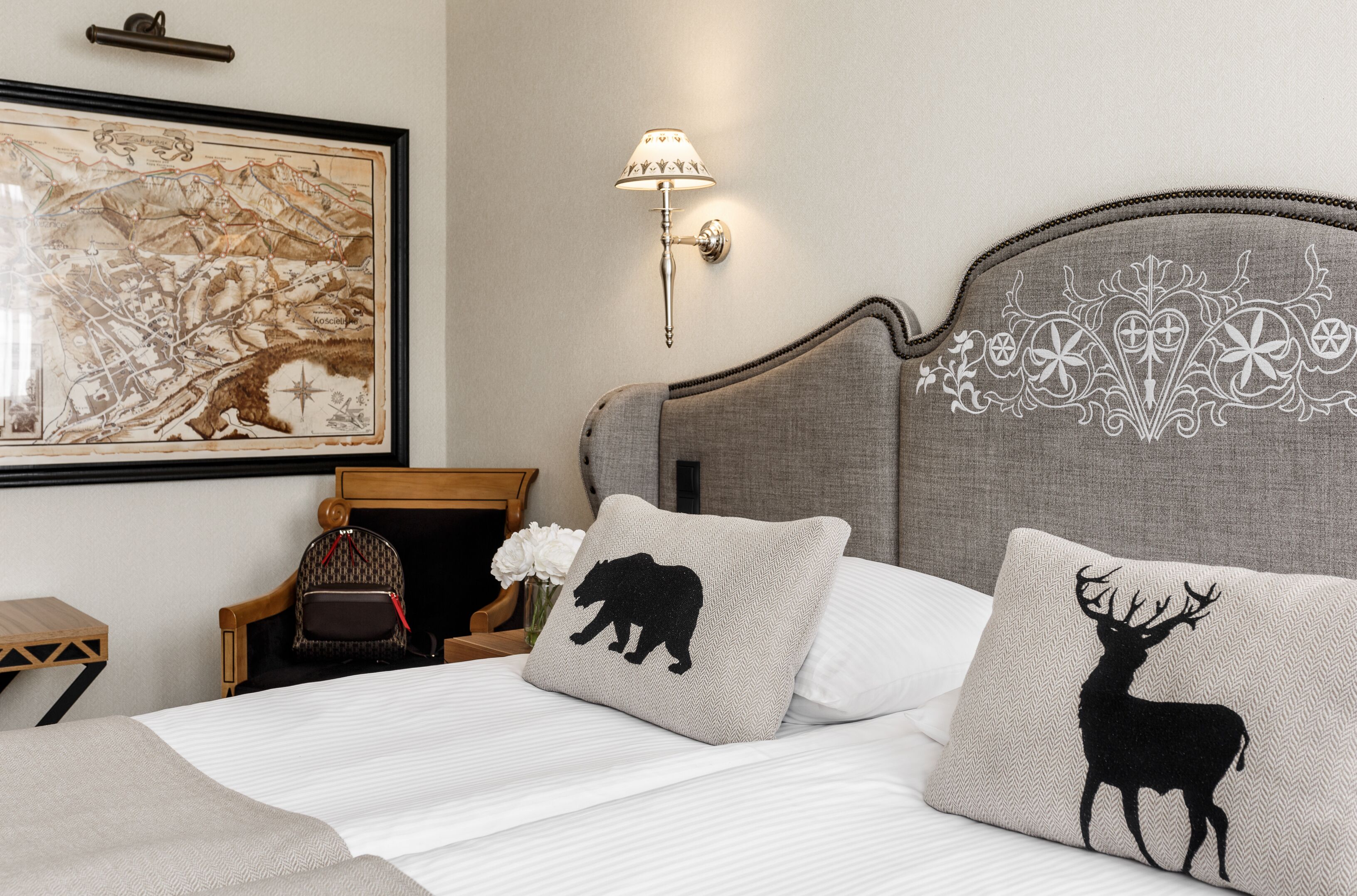 Una cama bien tendida con dos almohadas, una con silueta de oso y otra con silueta de ciervo, una lámpara de pared, un mapa enmarcado y una pequeña mesa de madera con una silla.
