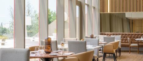 A modern restaurant interior with large windows, wooden slats on the ceiling, and a mix of grey and beige seating.