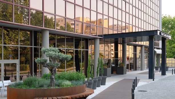 Una entrada de hotel moderna con fachada de vidrio, zona de estar al aire libre y jardín ajardinado.