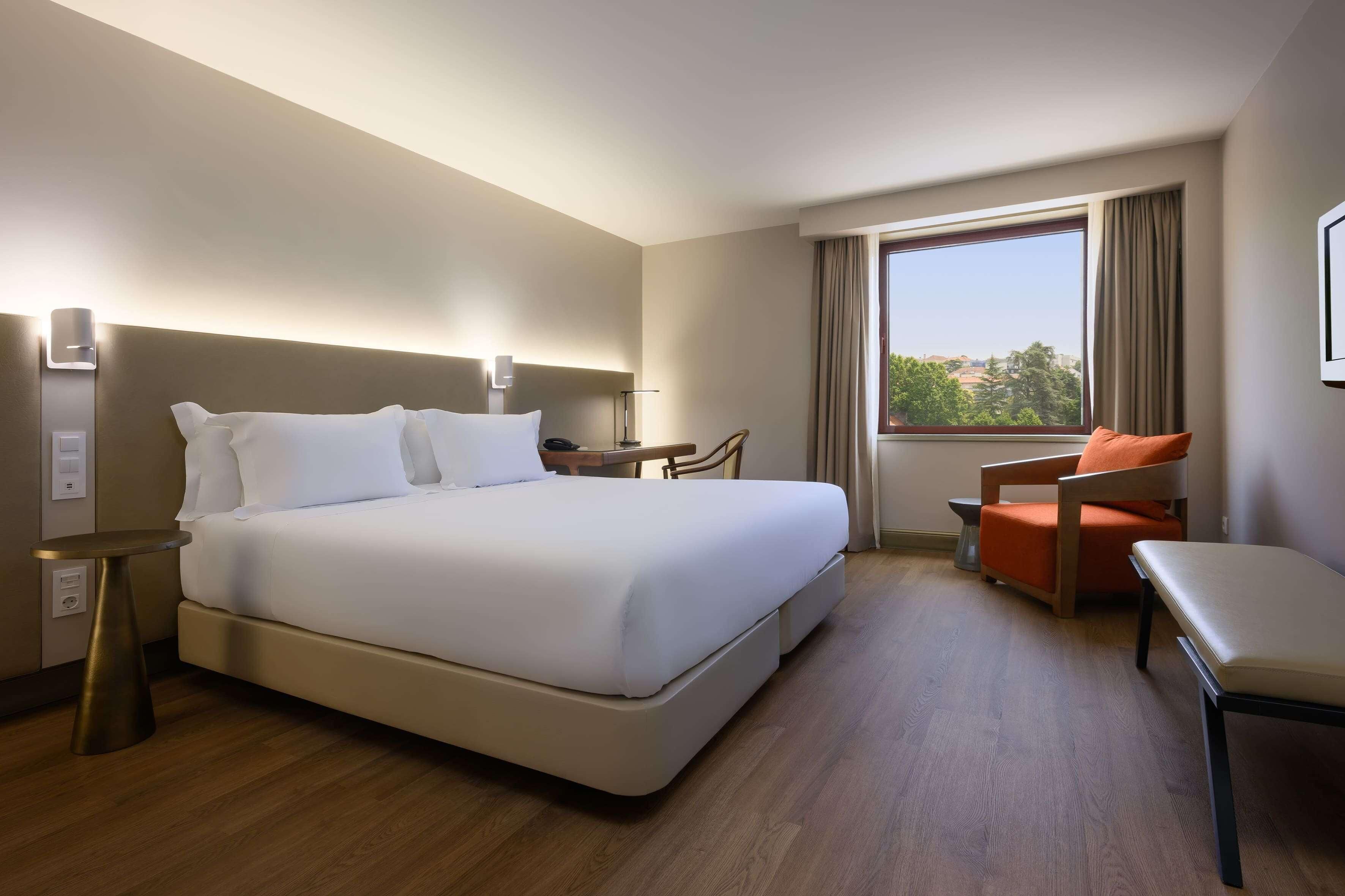 Habitación de hotel con cama, mesita de noche, silla y un ventanal que ofrece vistas.