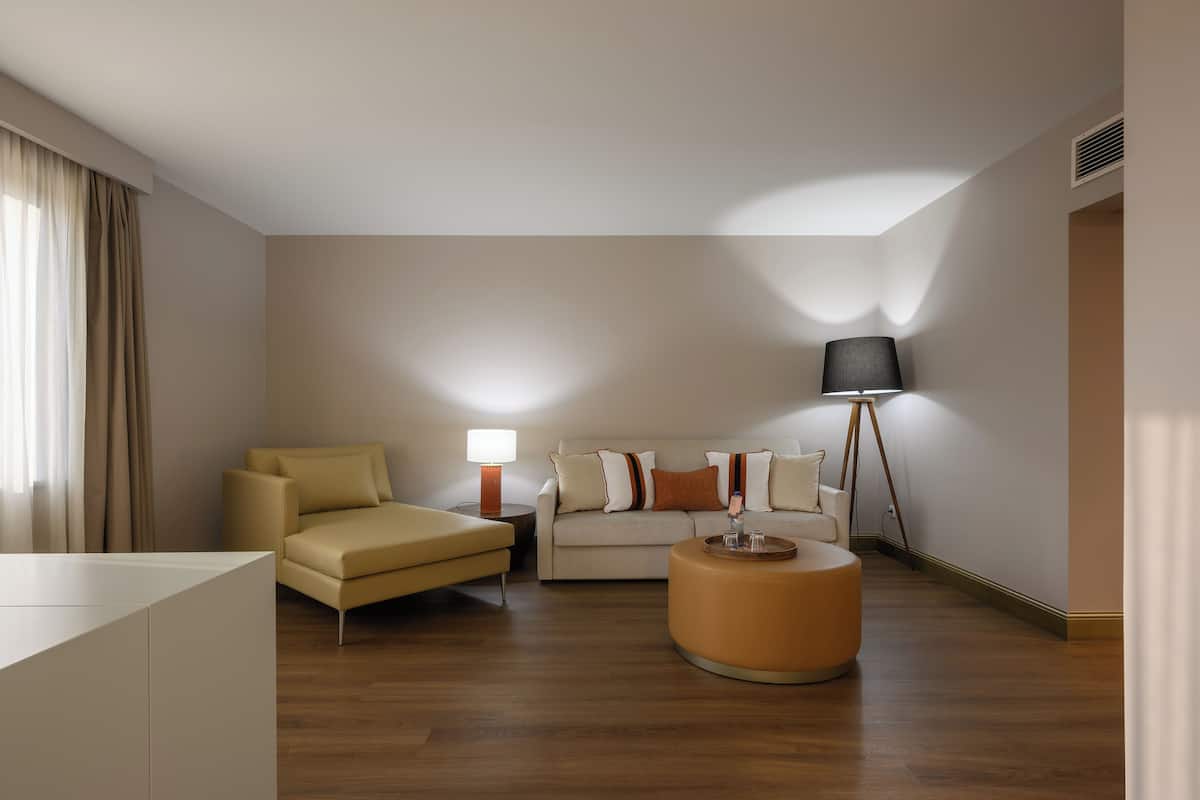 Una habitación de hotel moderna con un sofá, un otoman redondo, una mesita de noche con lámpara y una lámpara de pie.