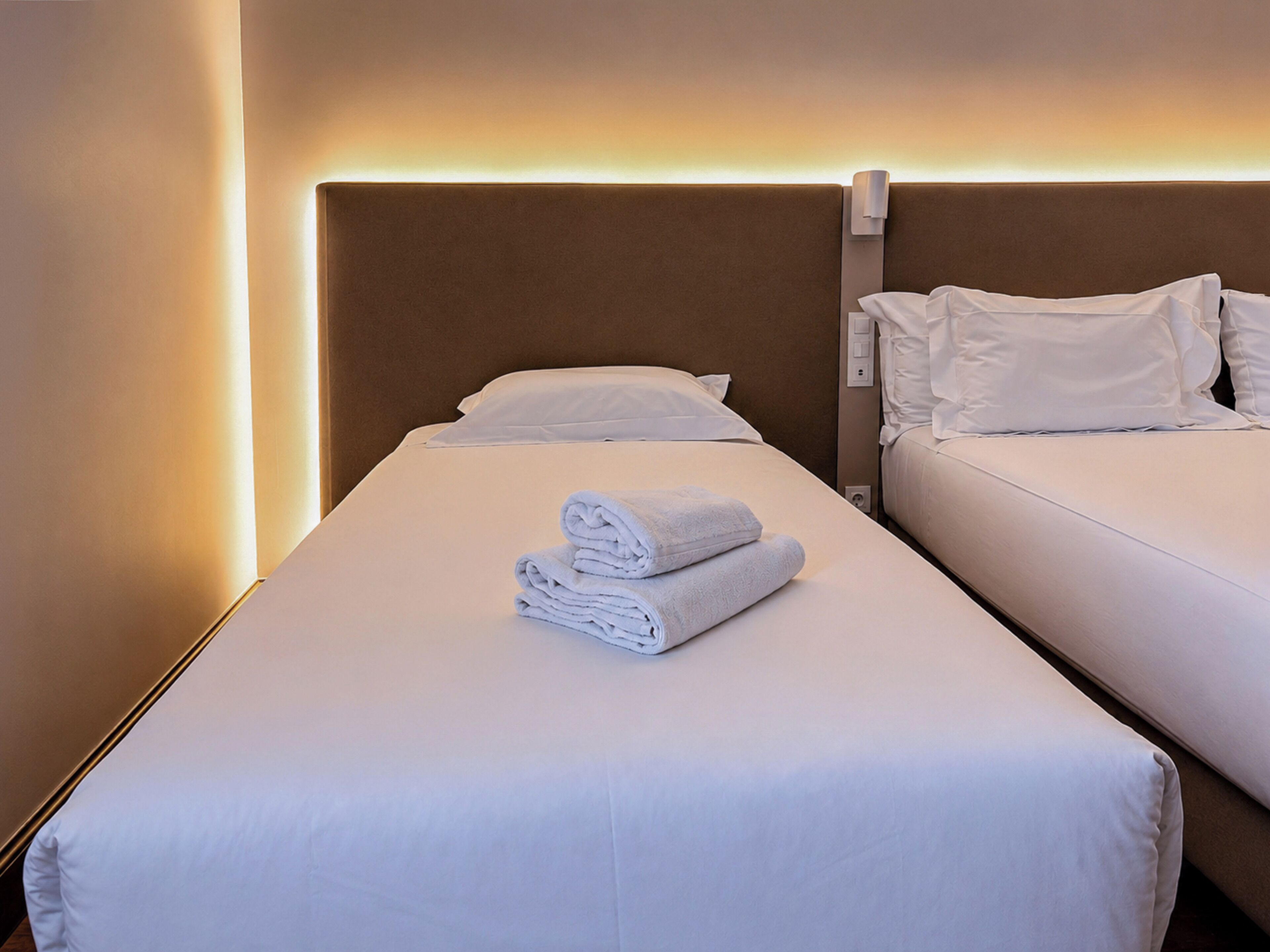 Habitación de hotel con dos camas, cada una con ropa blanca y almohadas, y una toalla doblada cuidadosamente sobre la cama en primer plano.