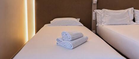 Habitación de hotel con dos camas, cada una con ropa blanca y almohadas, y una toalla doblada cuidadosamente sobre la cama en primer plano.