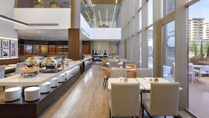 Un moderno vestíbulo de hotel con zona de buffet, asientos y amplias ventanas con vistas a la ciudad.