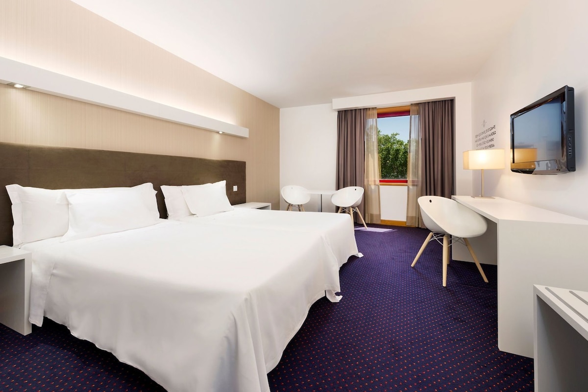A hotel room with a large bed, a desk, two chairs, a TV, and a window with curtains.