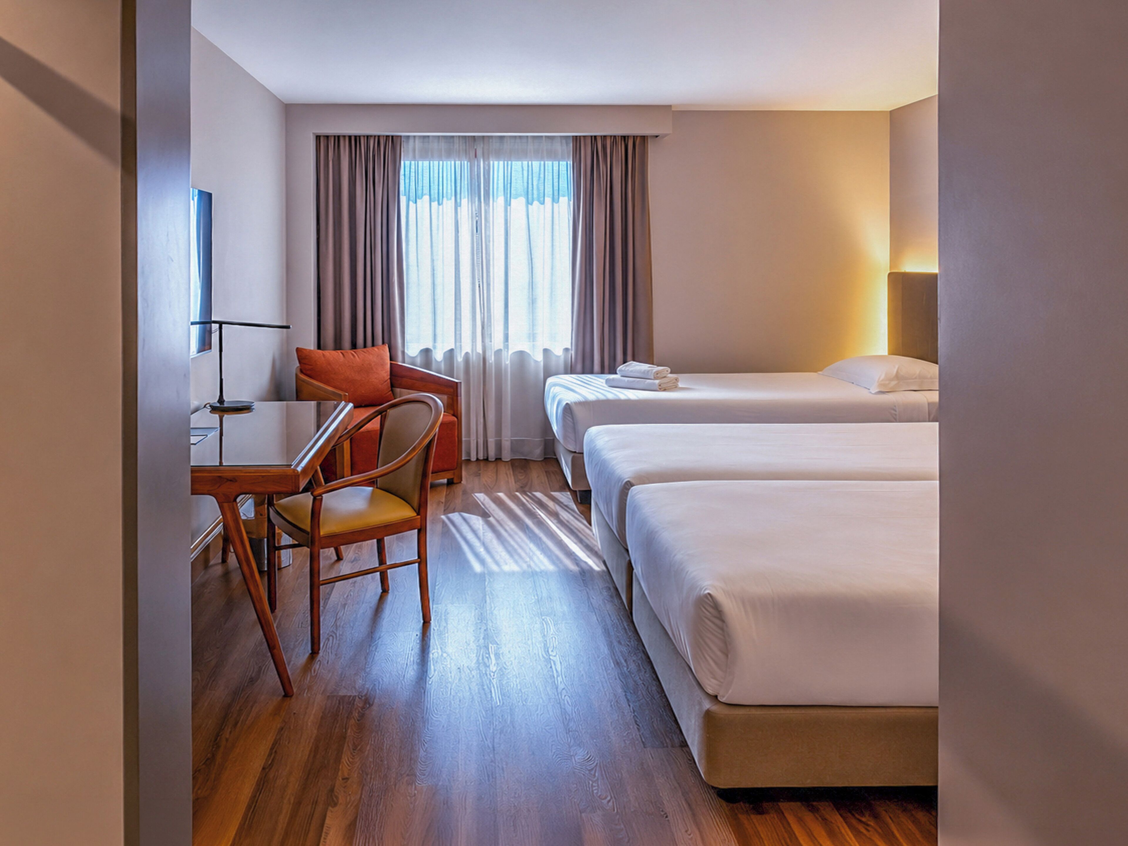 Een hotelkamer met een houten vloer, een bureau met stoel, twee enkele bedden en een raam met gordijnen.