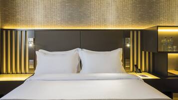 A modern hotel room with a large bed, two white pillows, a bedside table with a lamp, and a wall-mounted light fixture.