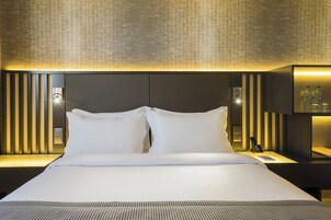 A modern hotel room with a large bed, two white pillows, a bedside table with a lamp, and a wall-mounted light fixture.