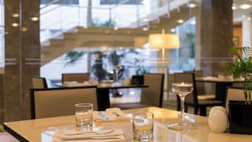 Mesa de comedor dispuesta con una vela, vasos y una planta en maceta en un moderno vestíbulo de hotel con vistas al interior.