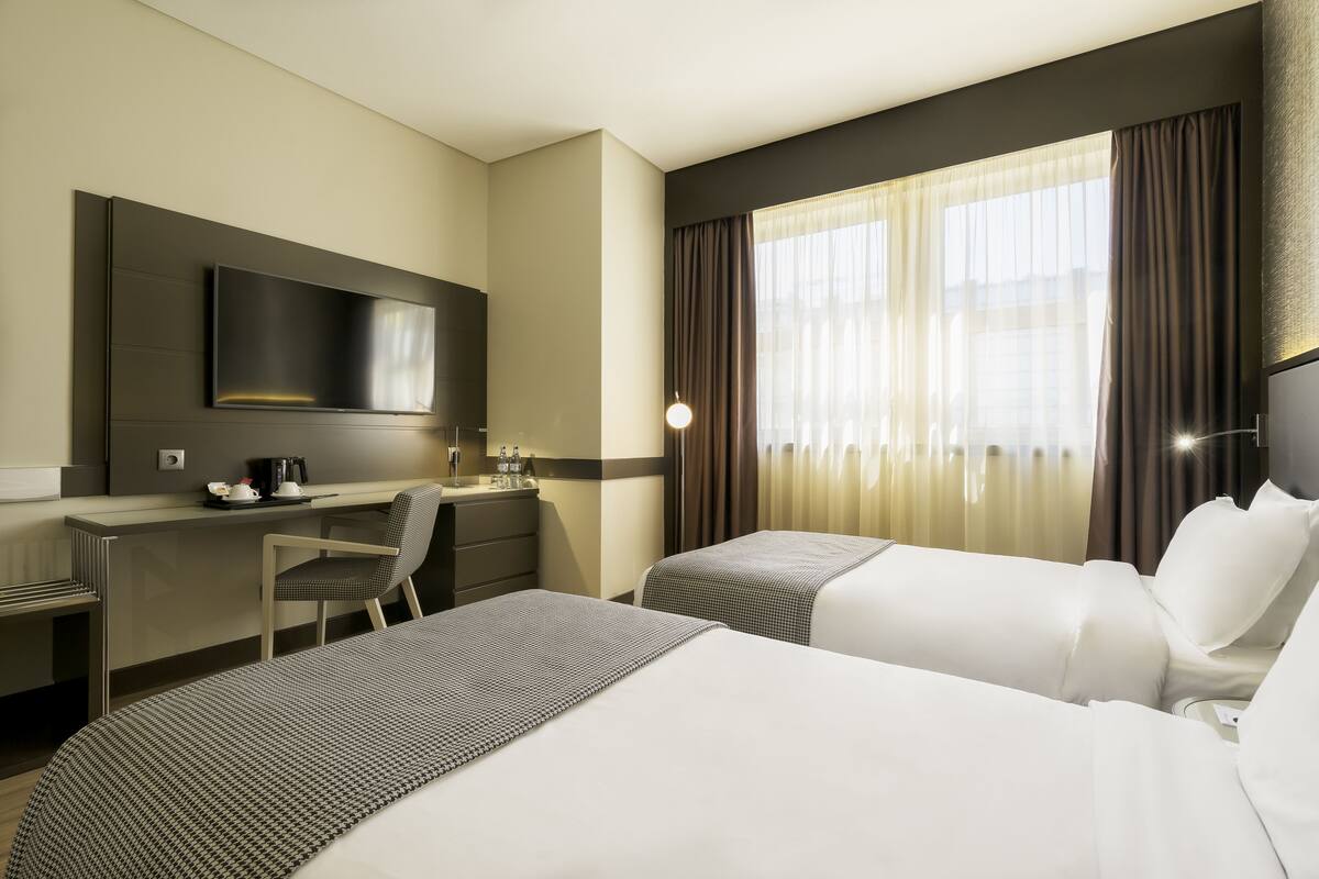 Ein Hotelzimmer mit zwei Betten, einem Schreibtisch, einem Stuhl, einem Fernseher und einem Fenster mit Vorhängen.