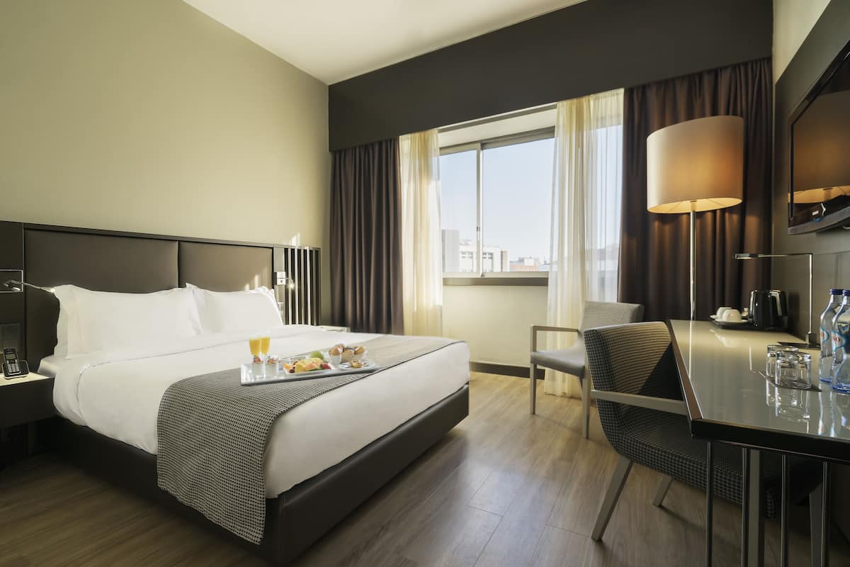 Ein Hotelzimmer mit einem großen Bett, einem Schreibtisch, einem Stuhl, einer Lampe und einem fenster mit Vorhängen.