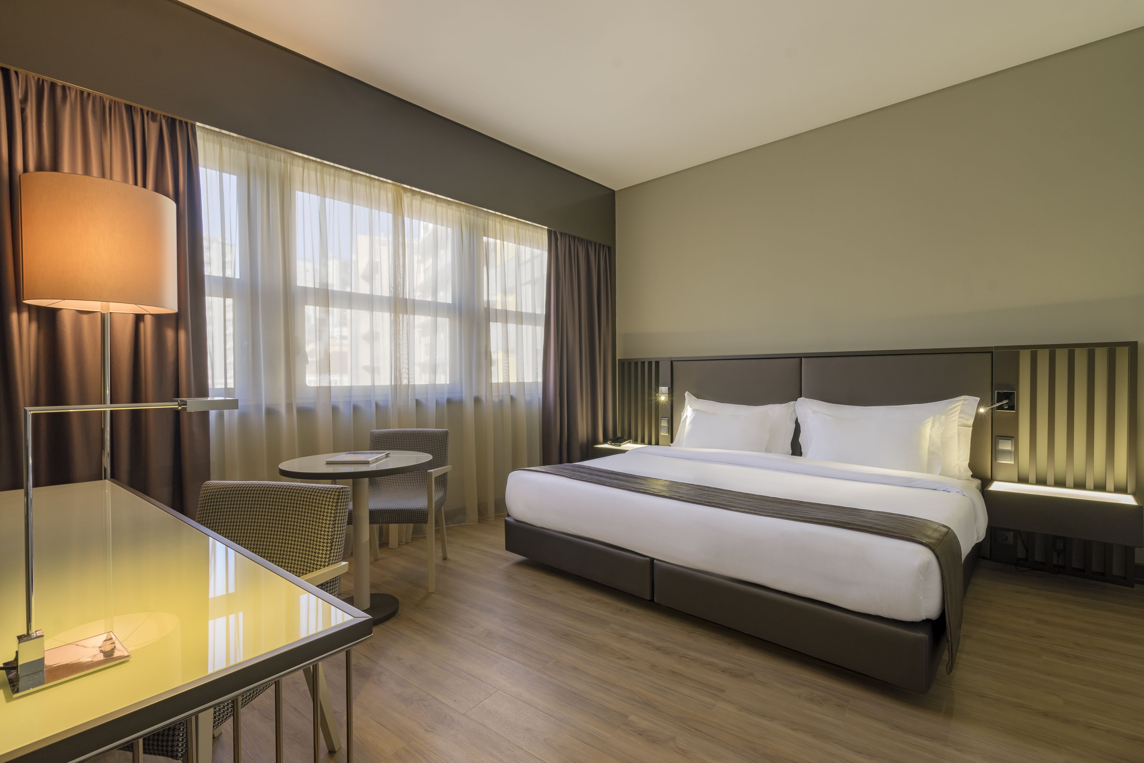 A hotel room with a bed, a desk, a chair, a lamp, and a window with curtains.