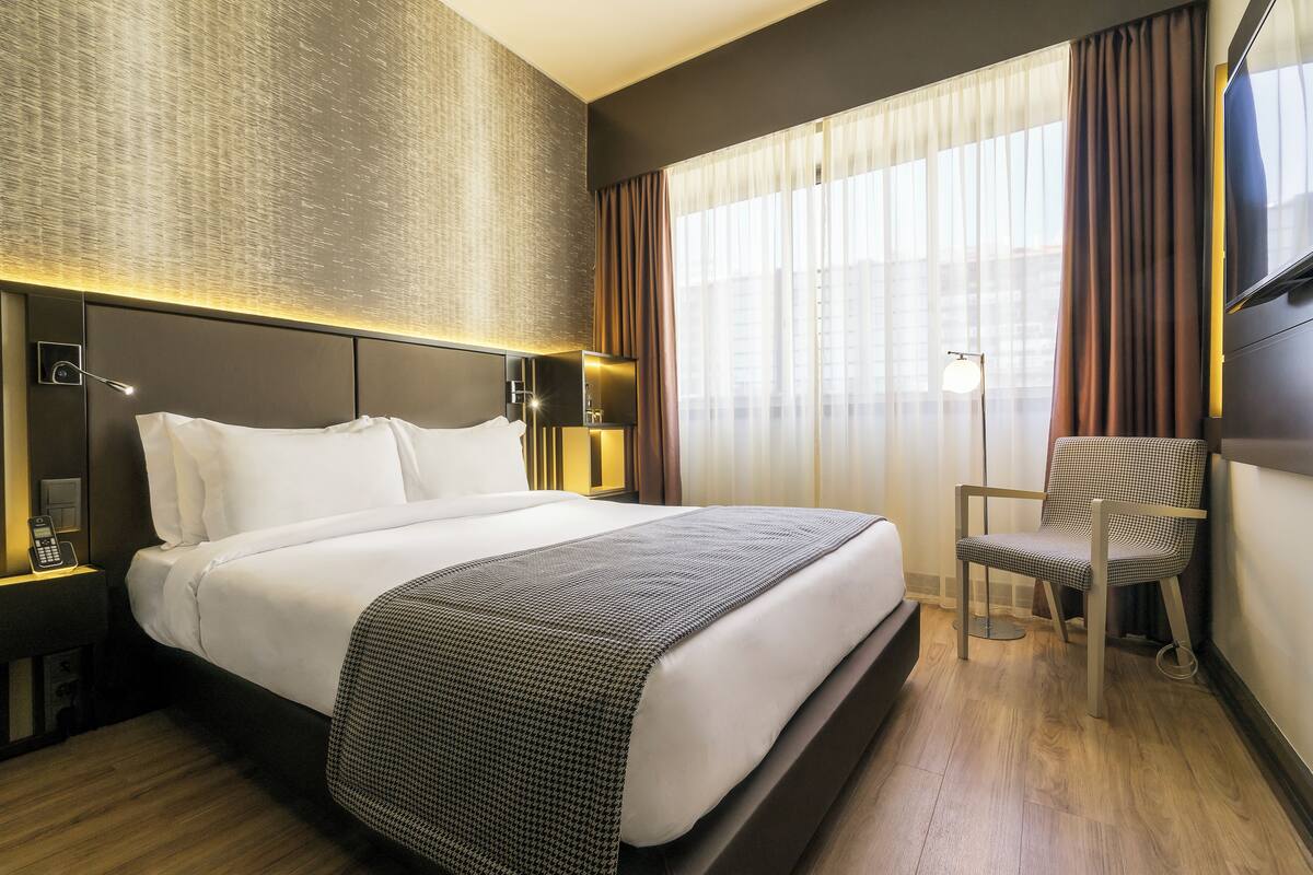 Ein Hotelzimmer mit einem großen Bett, einem Sessel, einem Schreibtisch mit Lampe sowie einem Fernseher an der Wand.