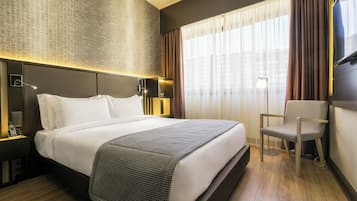 A hotel room with a large bed, a chair, a desk with a lamp, and a TV on the wall.