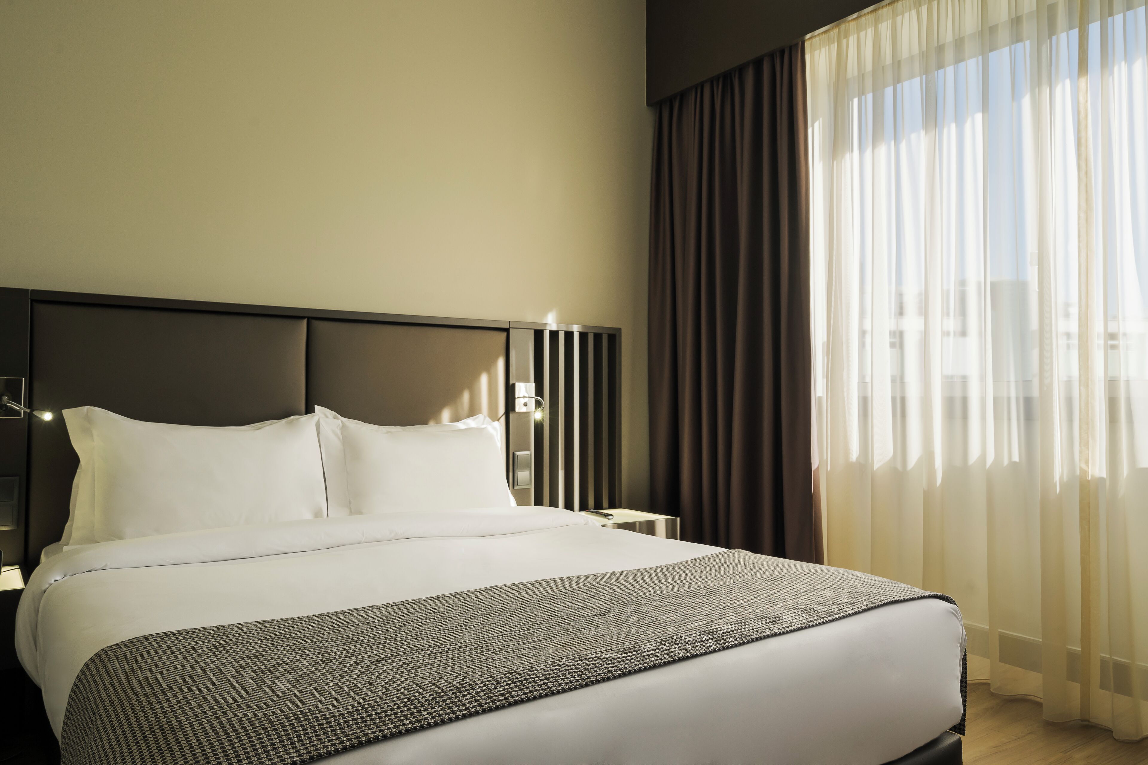 Cama bem arrumada com lençóis brancos e manta estampada, cabeceira moderna e vista da cidade através de cortinas translúcidas.