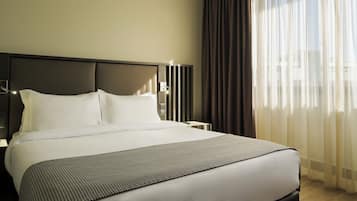 Cama bem arrumada com lençóis brancos e manta estampada, cabeceira moderna e vista da cidade através de cortinas translúcidas.