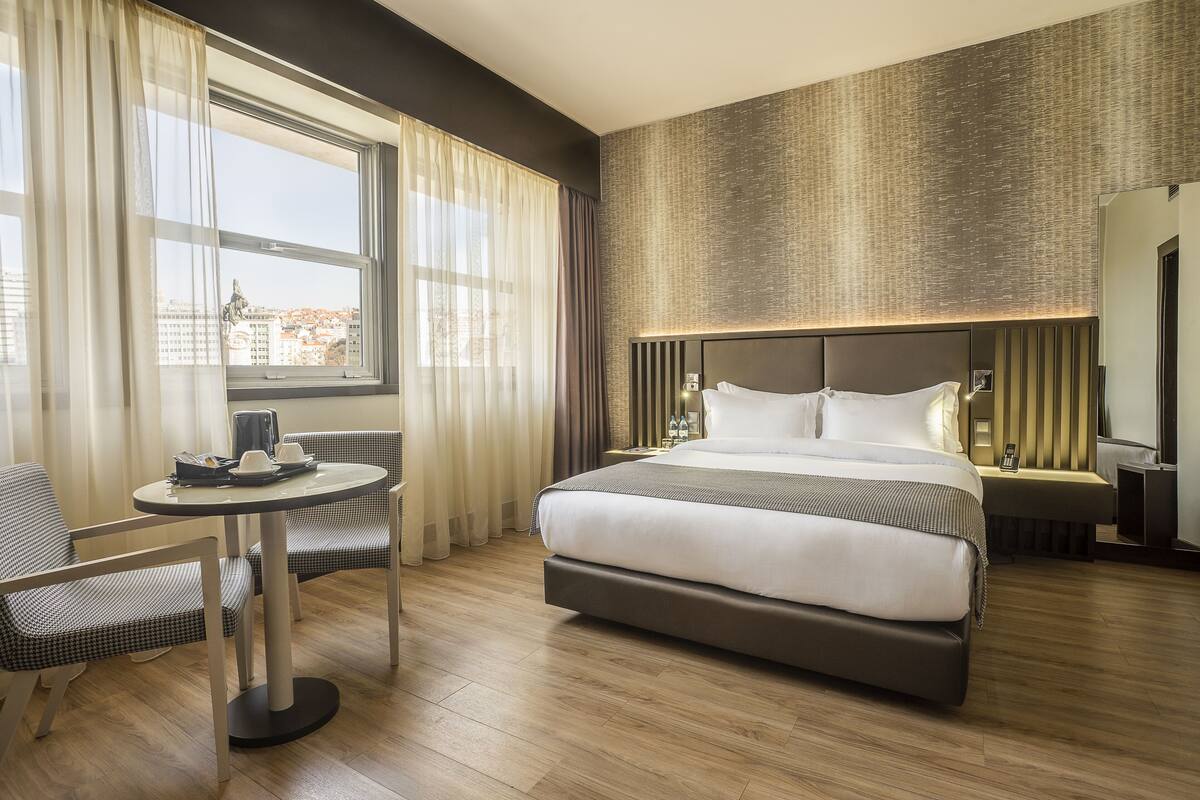Ein Hotelzimmer mit einem großen Bett, einem kleinen runden Tisch, zwei Stühlen und Blick auf die Stadt durch das Fenster.