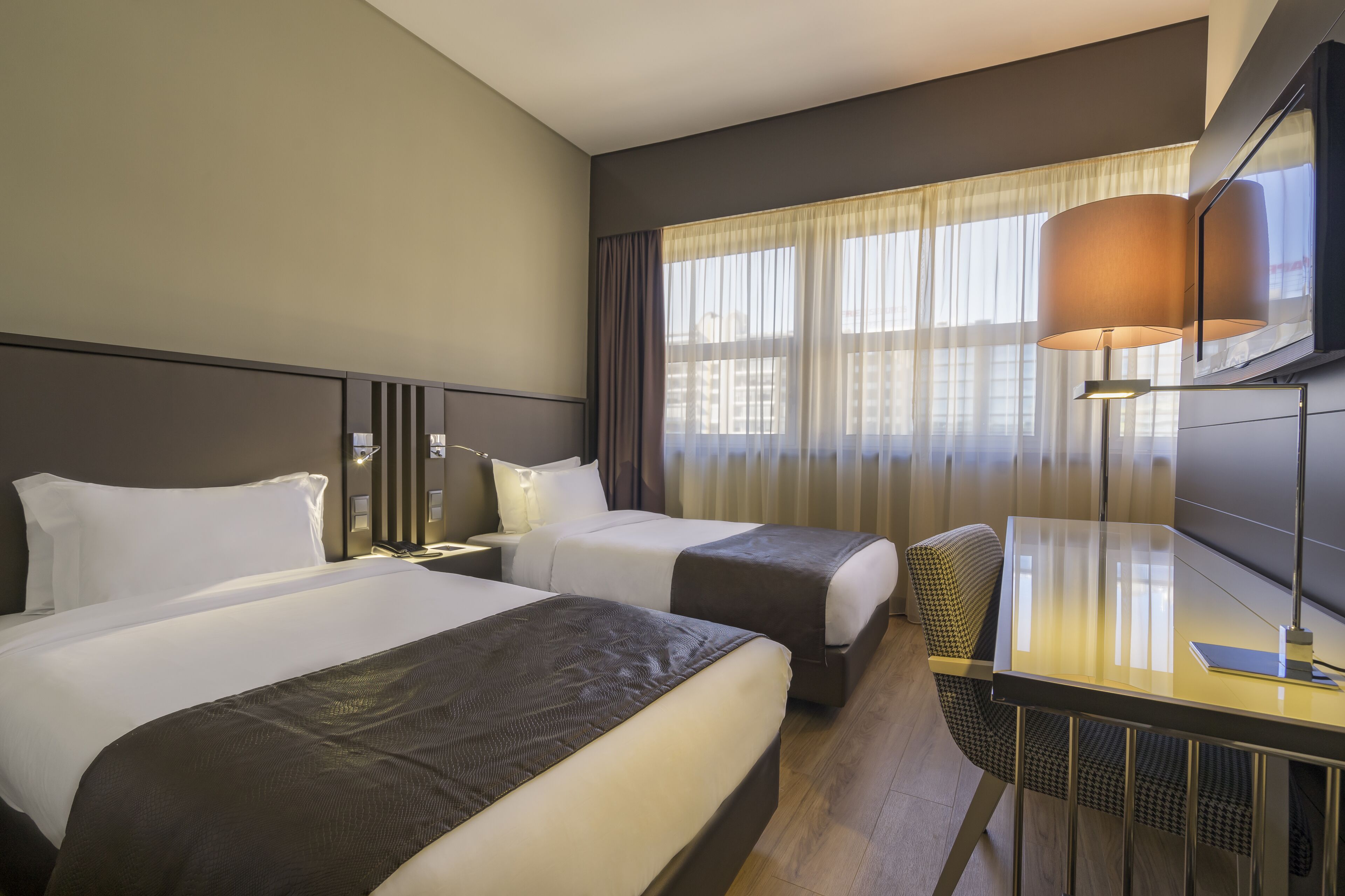 Hotellihuone, jossa on kaksi sänkyä, työpöytä, tuoli, televisio ja ikkuna, jossa on verhot.
