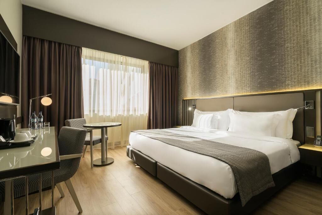 Habitación de hotel con cama, mesita de noche, lámpara, silla y vistas al exterior a través de la ventana.