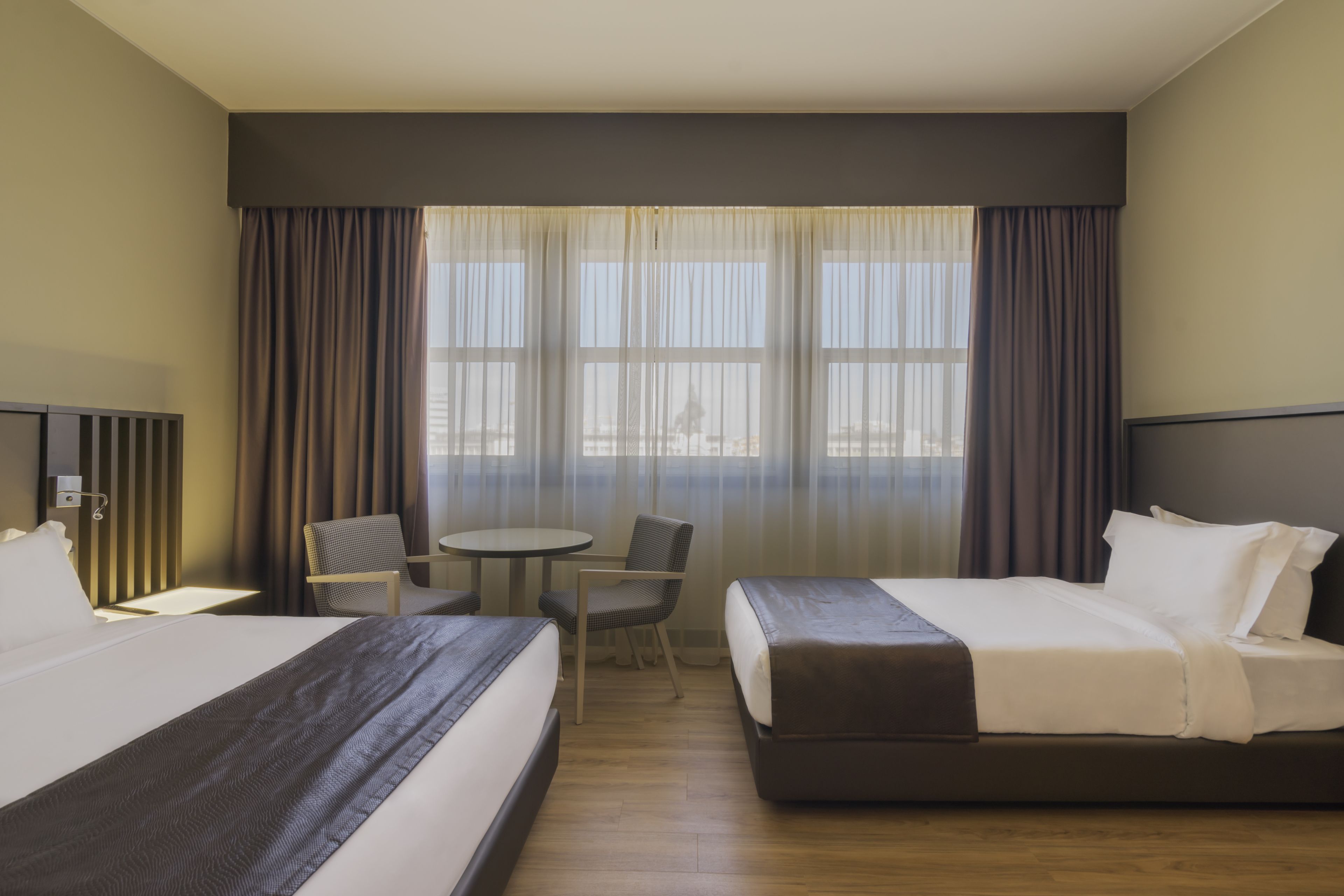 A hotel room with two beds, a small table, two chairs, and a view of the city through large windows.