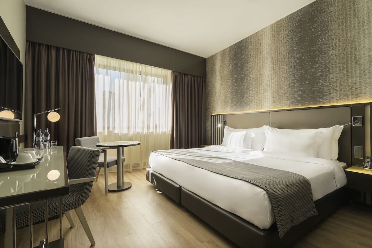Ein modernes Hotelzimmer mit einem großen Bett, einem Schreibtisch mit Stuhl, einem kleinen Tisch und einem großen Fenster mit Vorhängen.