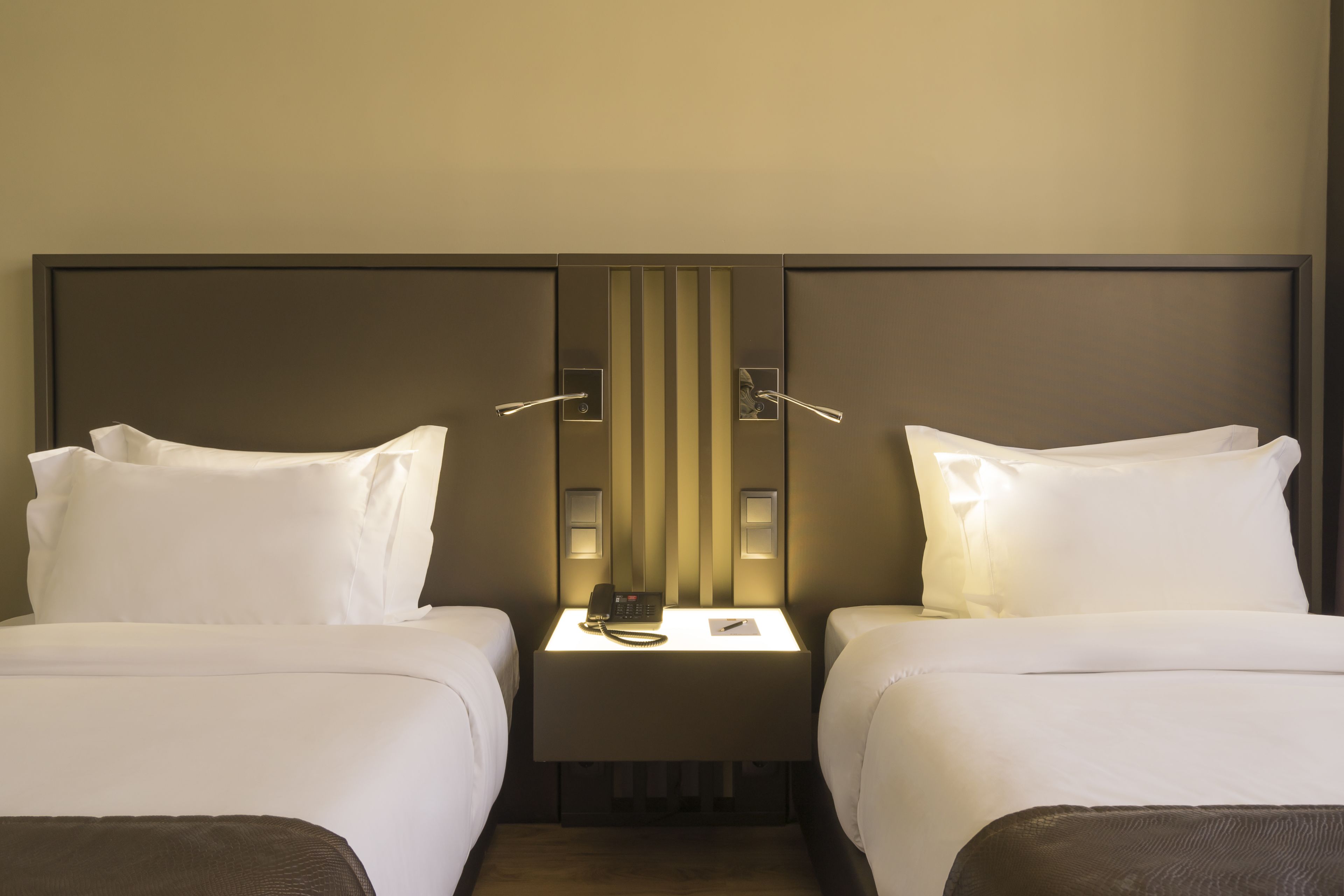 A hotel room with two beds, a bedside table with a phone, and a wall-mounted light.