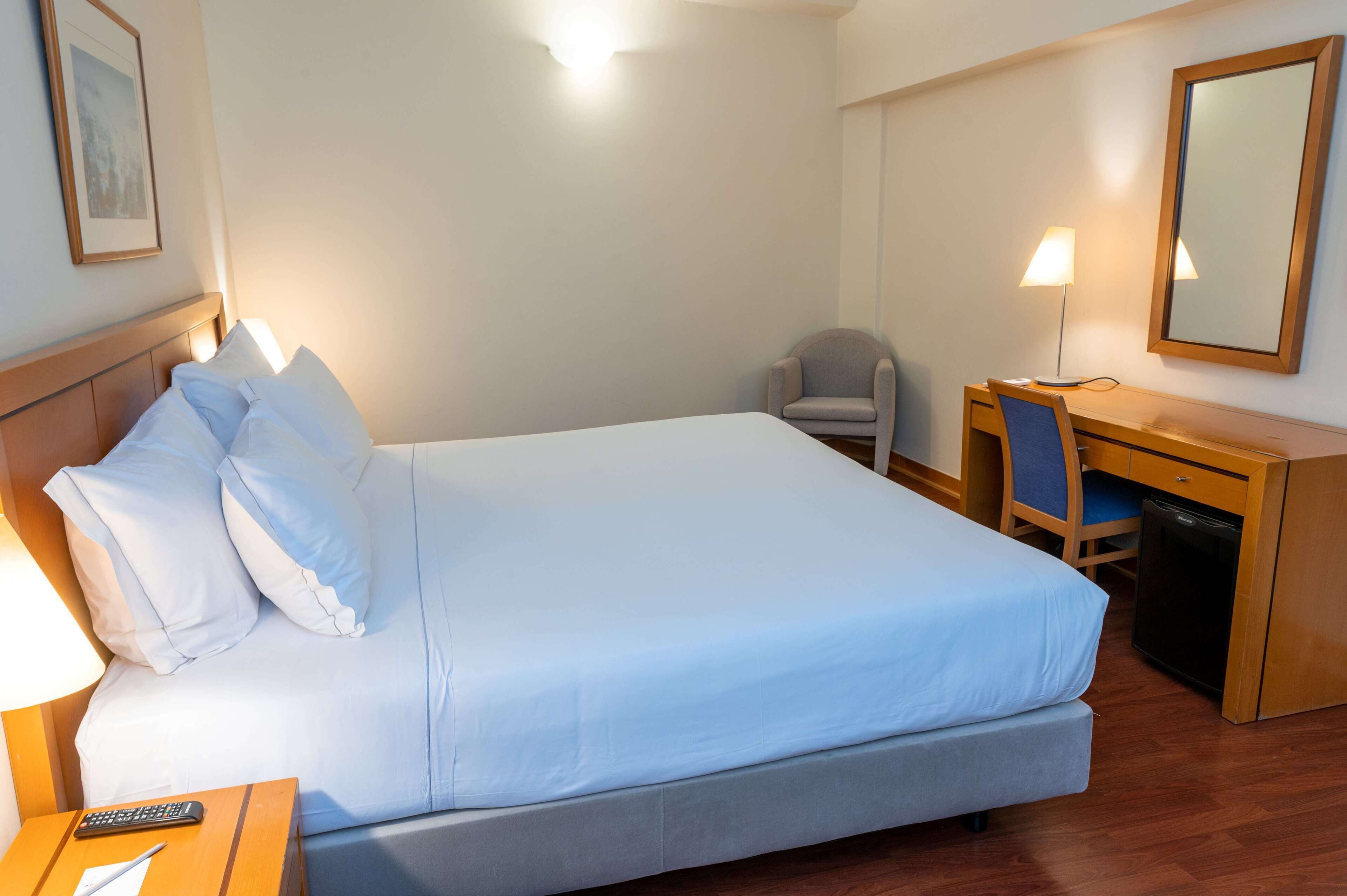 A hotel room with a bed, a desk, a chair, a lamp, and a small cabinet.