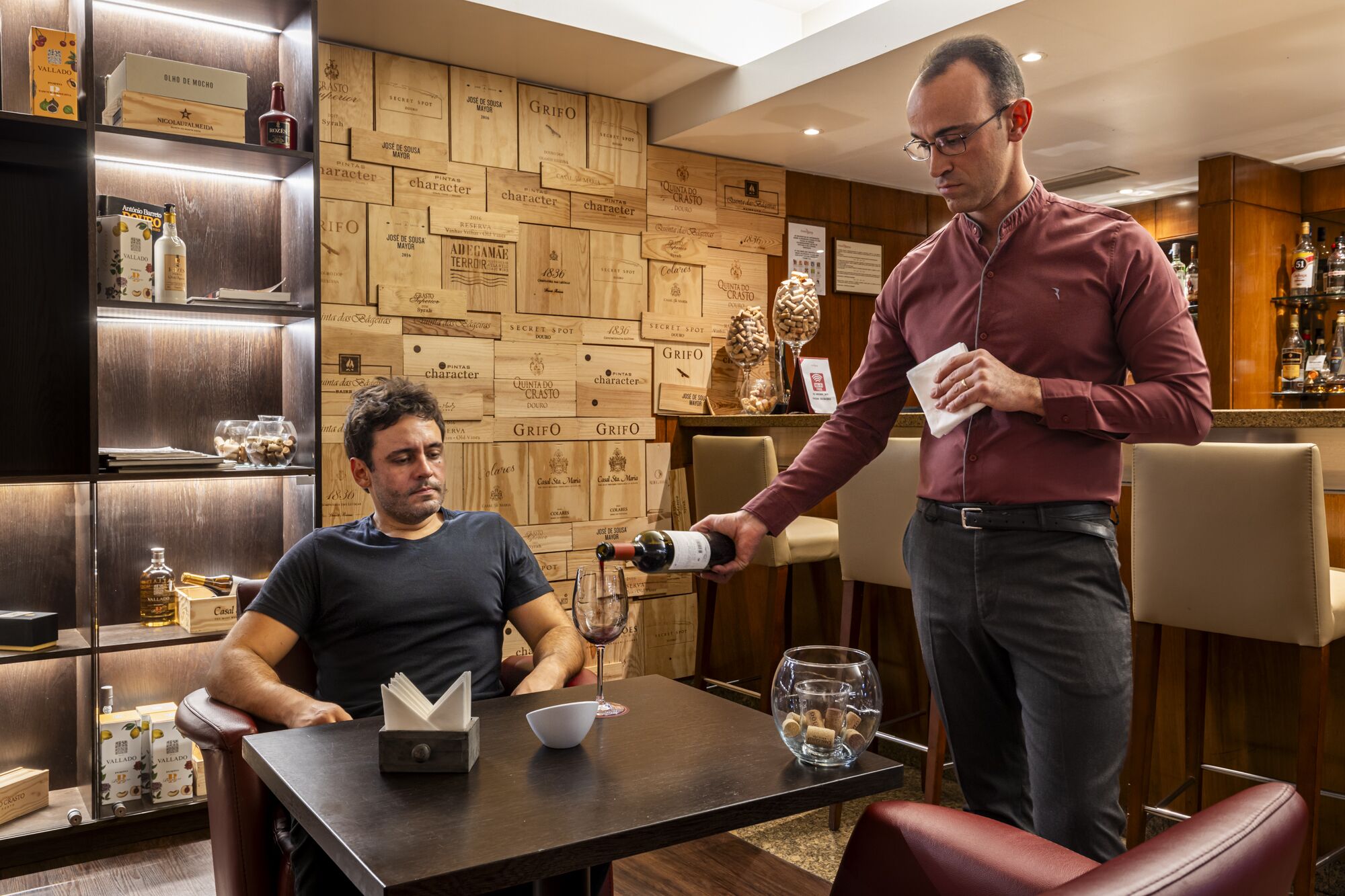 A man pouring wine into a glass at a bar with wooden wall panels and various bottles and boxes.