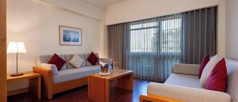 Una habitación de hotel moderna con piso de madera, un sofá, una cama, una mesita de centro y un ventanal con cortinas.