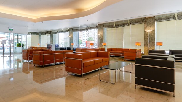 A spacious lobby with modern orange sofas, a black sofa, and a glass coffee table. Large windows with blinds provide natural light.