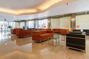 A spacious lobby with modern orange sofas, a black sofa, and a glass coffee table. Large windows with blinds provide natural light.